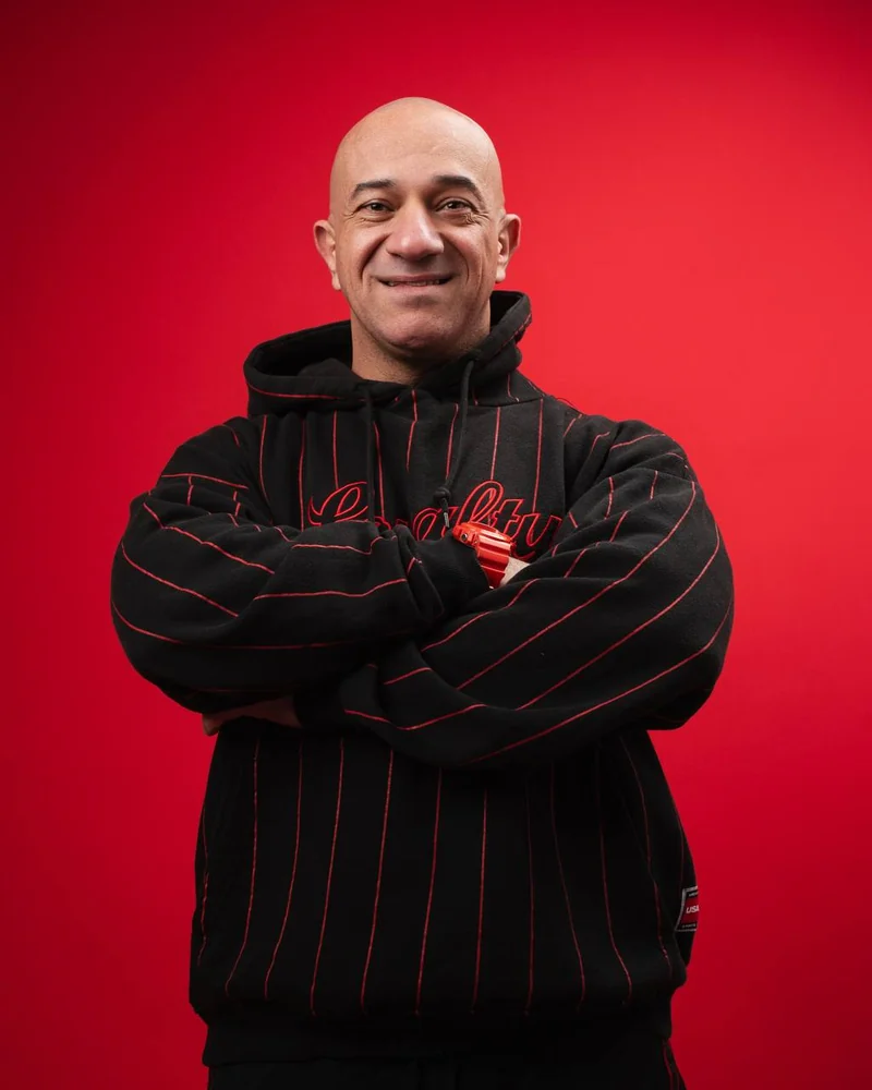Akmal Kadri with arms crossed in a black and red hoodie against a red background